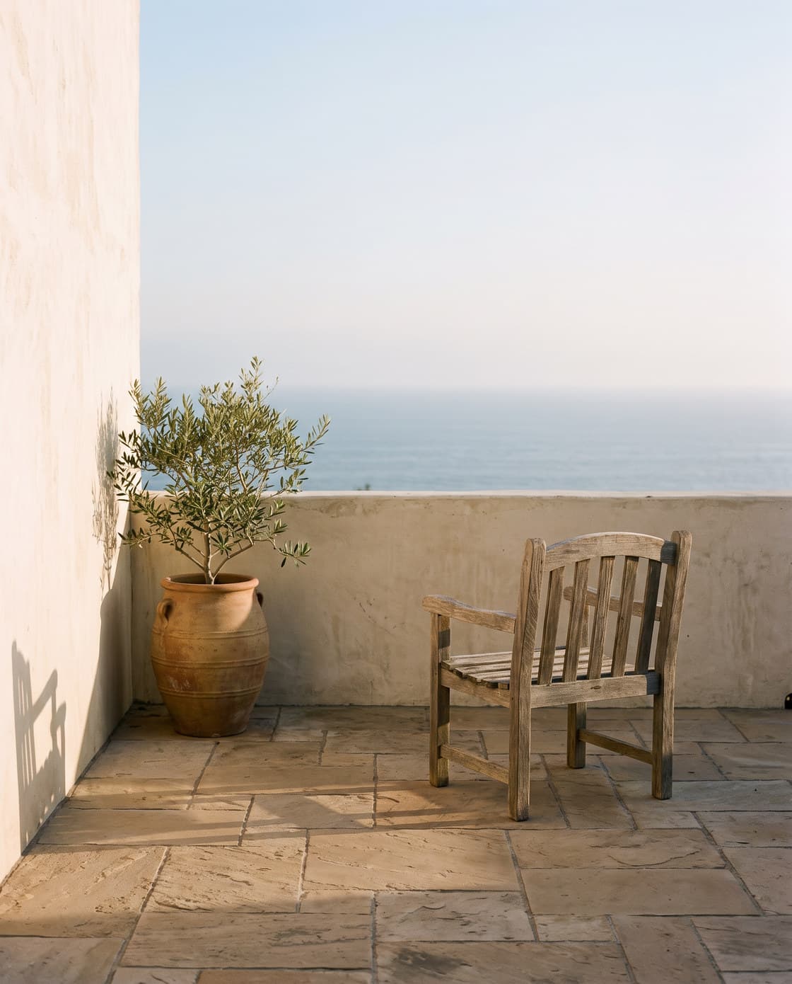 A single weathered wooden chair on a quiet stone terrace above the sea, beside an olive tree in a terracotta pot.