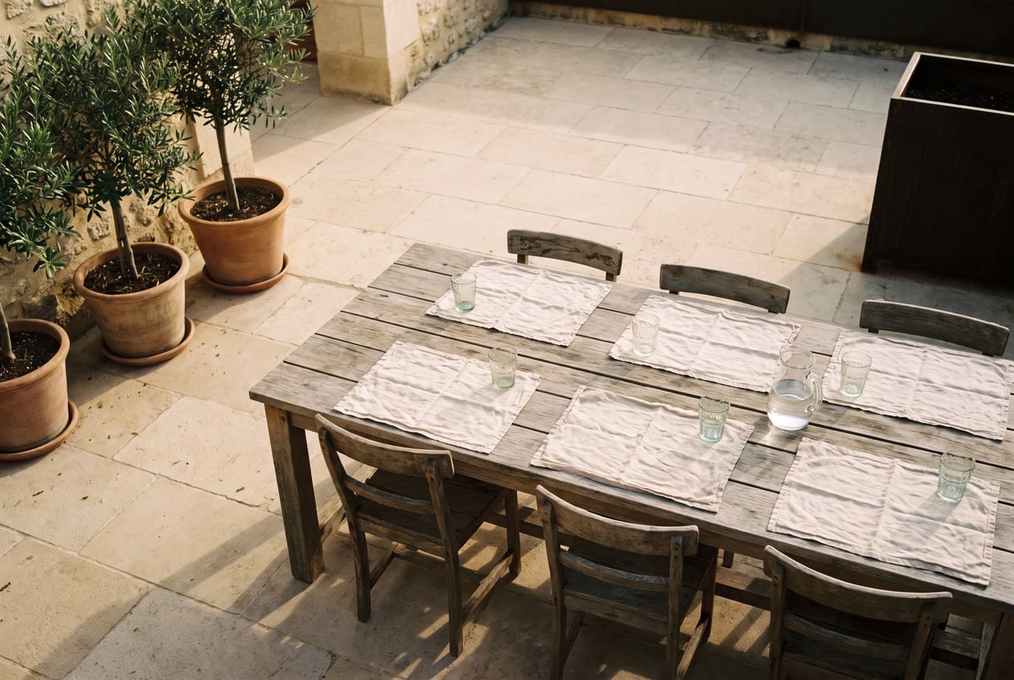 An outdoor table laid for six on a sunlit stone terrace, before anyone has arrived.