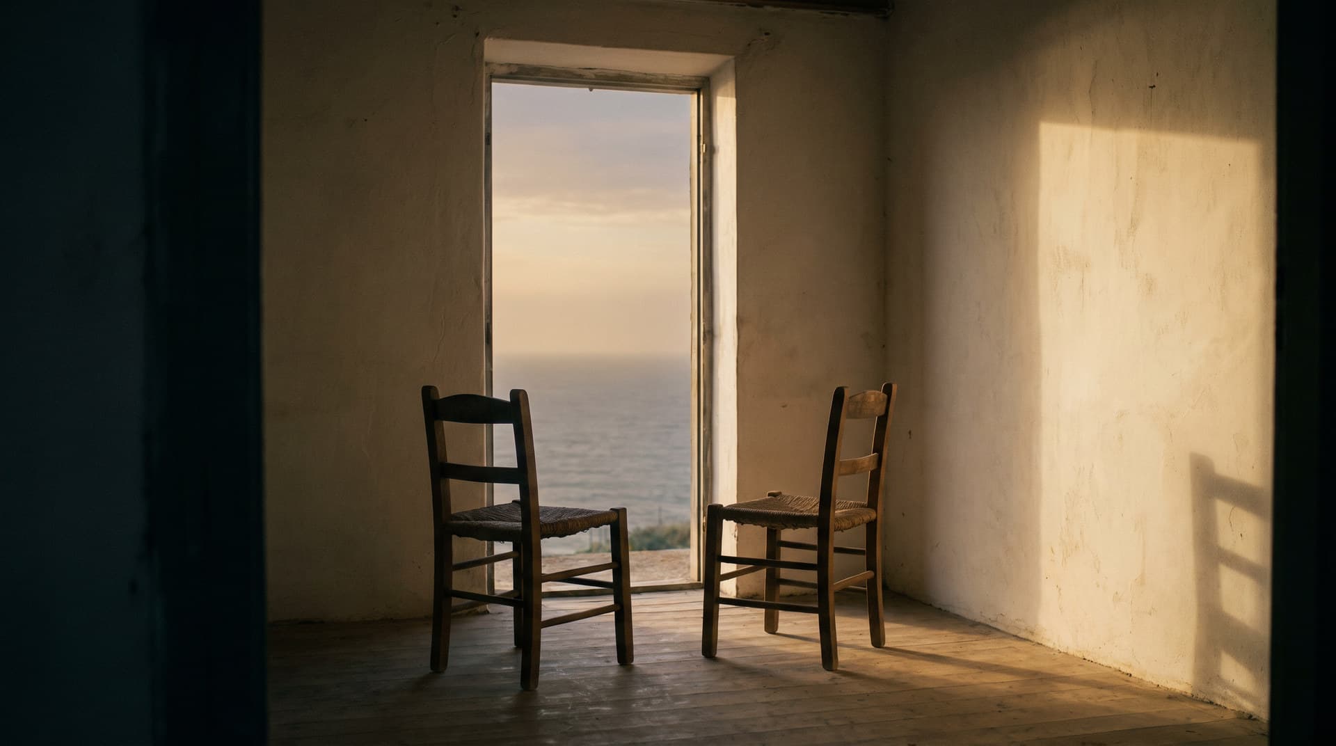 A quiet room opening onto the sea through a tall unglazed opening, with two simple chairs facing each other in late light.